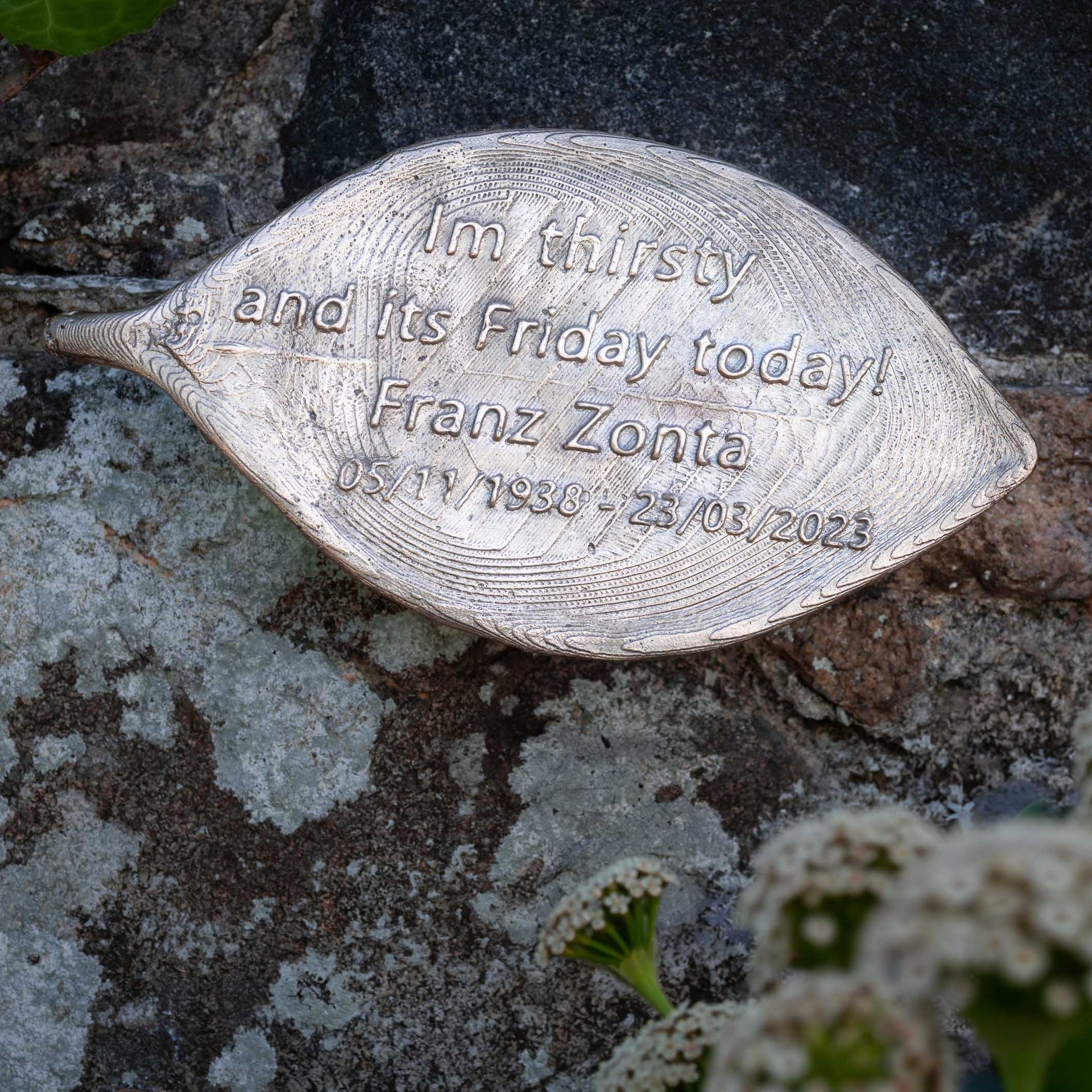 Bronze leaf-shaped object with engraved text on a stone surface with plants
